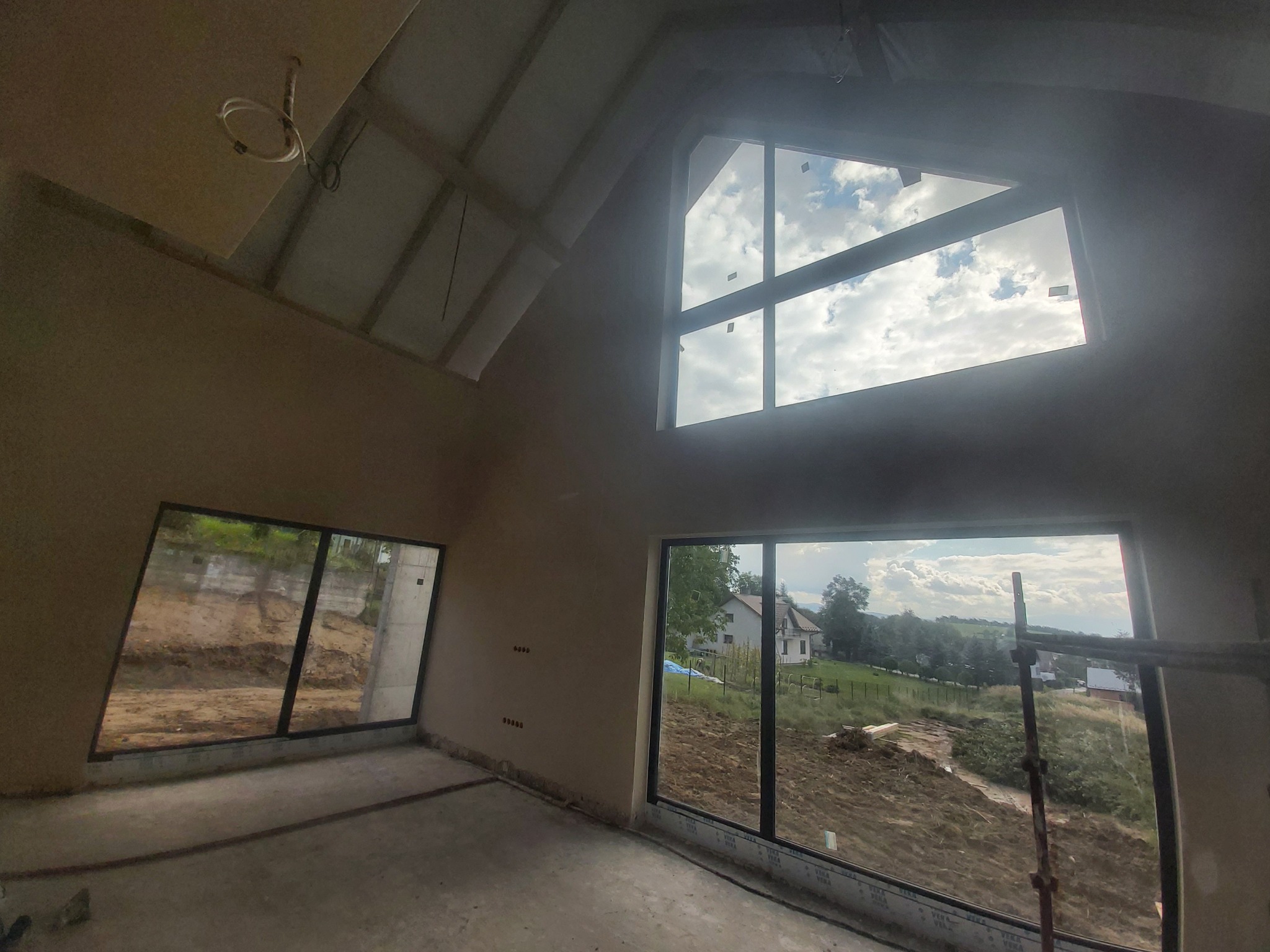 Wnętrze poddasza w stanie deweloperskim po nałożeniu jasnych tynków z widocznymi oknami i elementami instalacji elektrycznej.