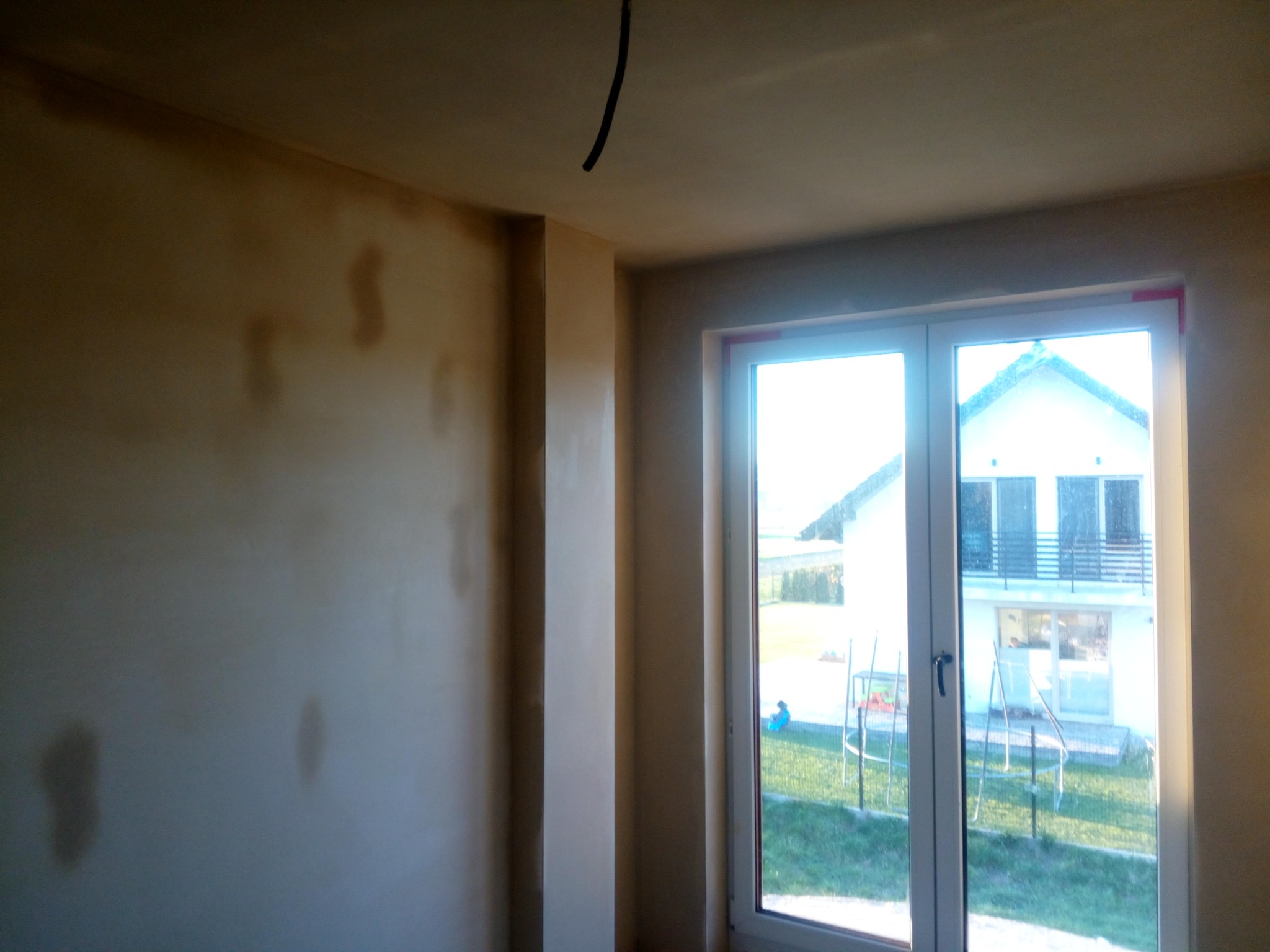 Pokój w trakcie prac wykończeniowych: ściany częściowo pokryte tynkiem, widoczne plamy wilgoci, okno z widokiem na dom i ogród.