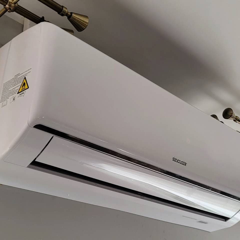 Biały klimatyzator Sinclair zamontowany na ścianie pod sufitem, widoczna lampa w tle.