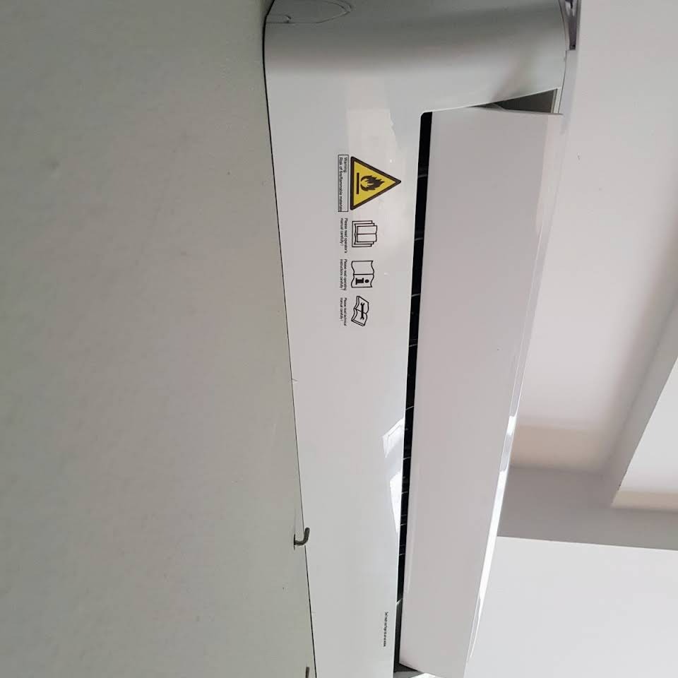 Biała jednostka wewnętrzna klimatyzatora zamontowana na ścianie, widoczne naklejki ostrzegawcze i informacyjne po lewej stronie.