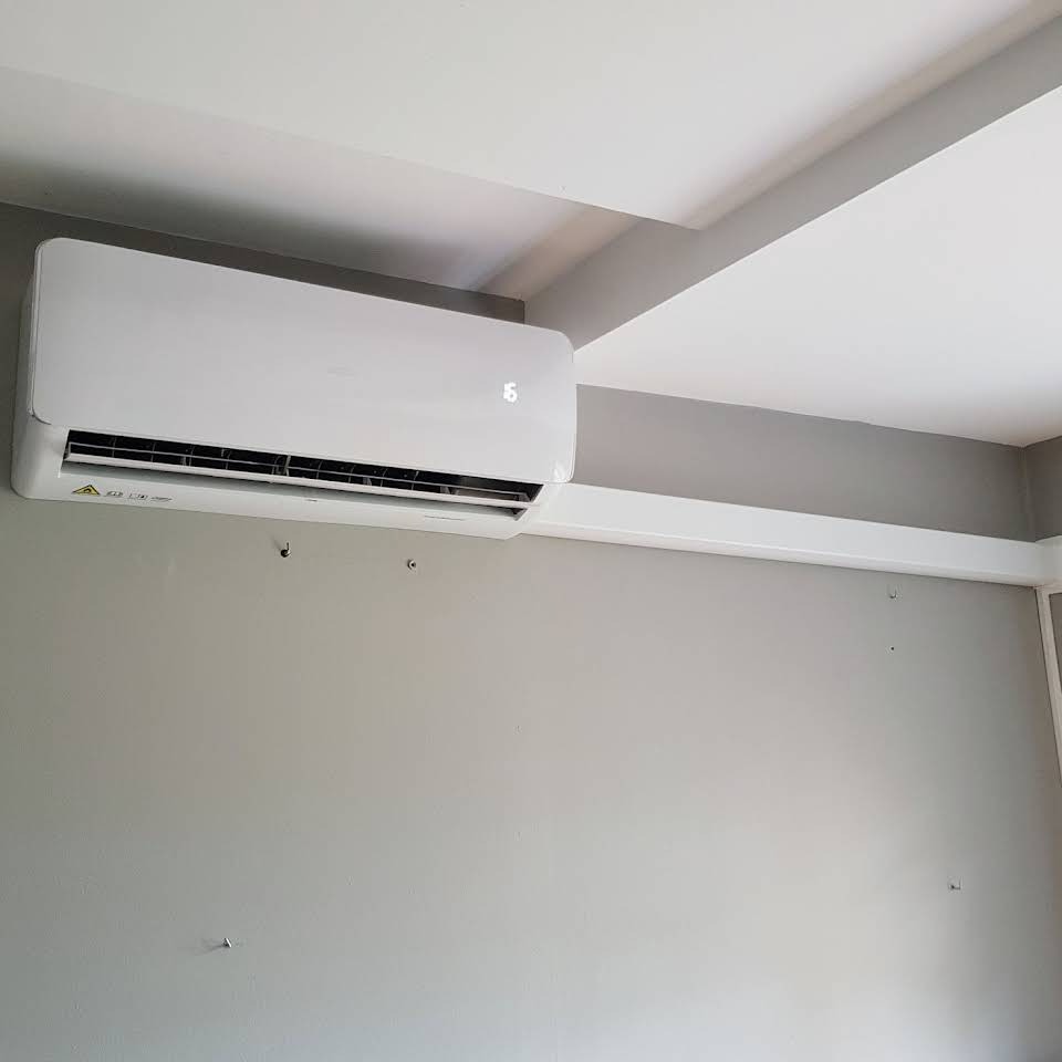 Biały klimatyzator ścienny z wyświetlaczem cyfrowym zamontowany na szarej ścianie, widoczne ślady po wcześniejszych mocowaniach i listwa maskująca instalację.