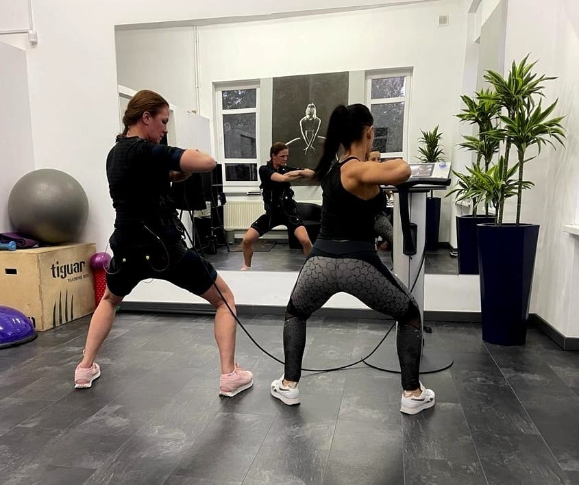 Trzy osoby ćwiczą w strojach do elektrostymulacji w sali fitness z lustrami, widoczne rośliny doniczkowe i sprzęt sportowy Tiguar.