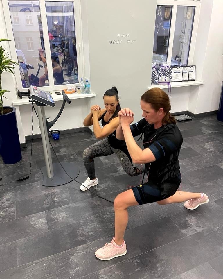 Dwie kobiety w strojach do elektrostymulacji mięśni wykonują ćwiczenia: przysiady i wykroki w jasnym pomieszczeniu z dużym oknem i napisem 'EMS Body Fit' na ścianie.