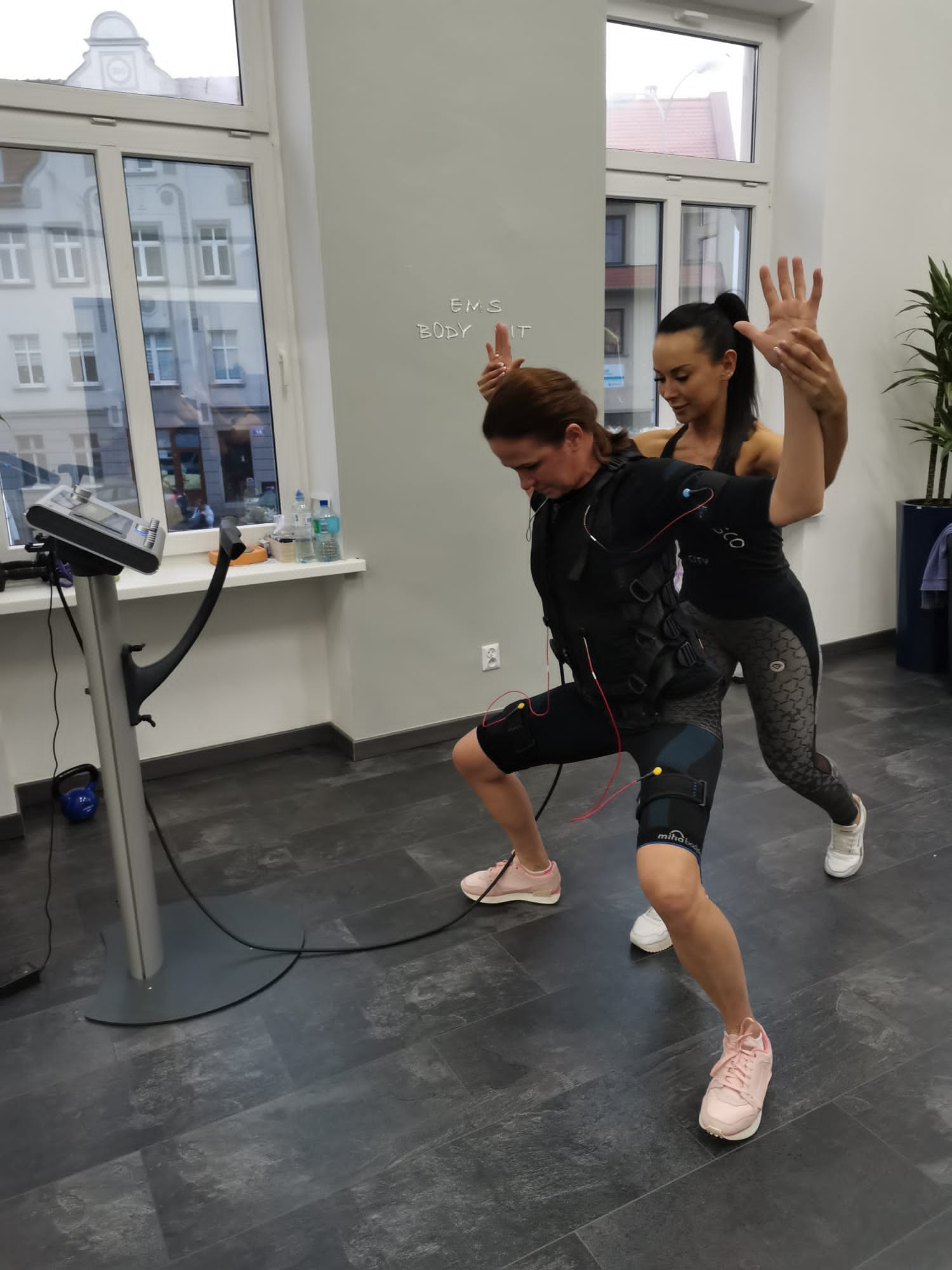 Kobieta w kombinezonie EMS ćwiczy pod okiem trenerki w jasnym pomieszczeniu z dużym oknem.