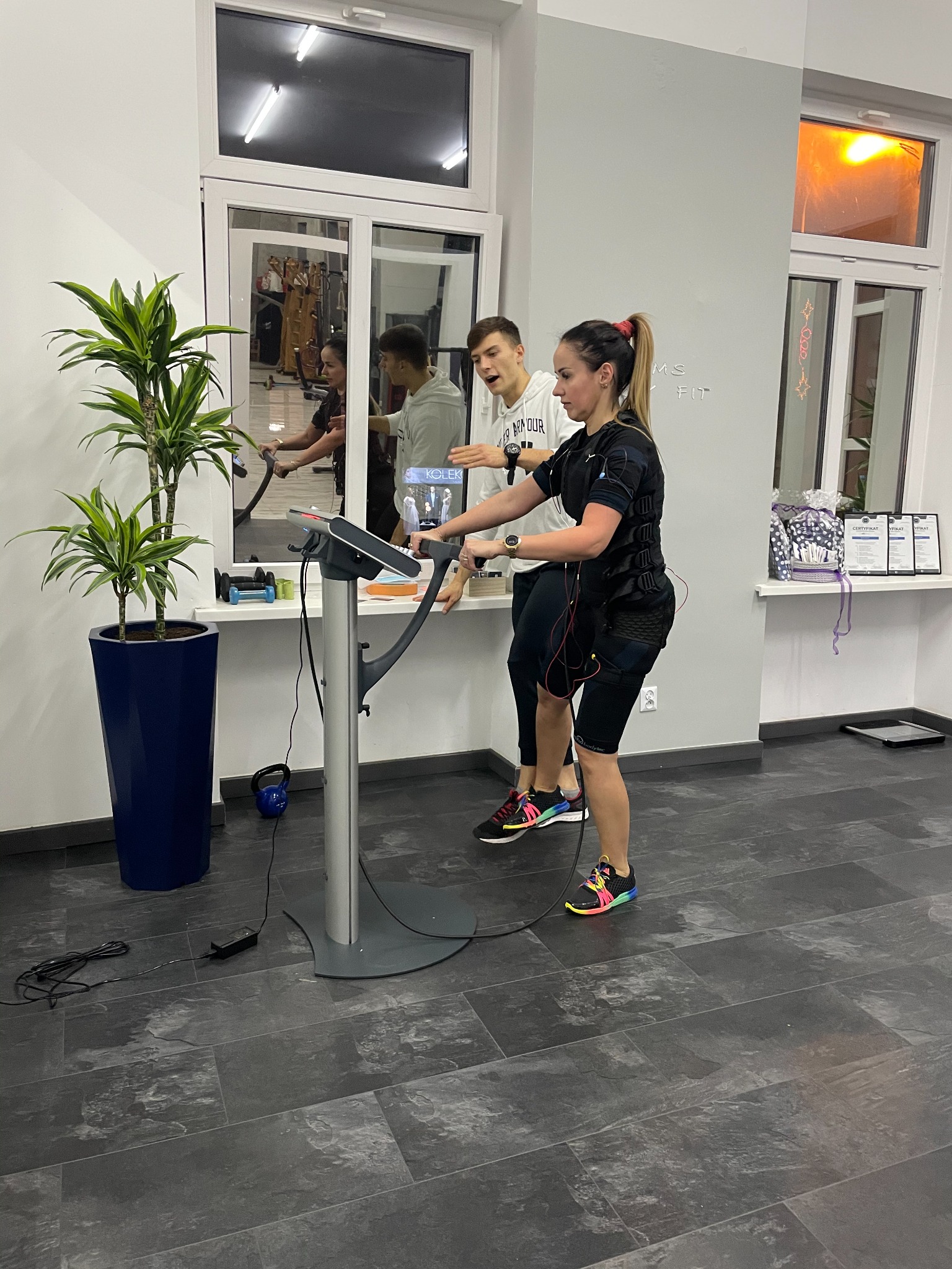 Kobieta w kombinezonie EMS ćwiczy na platformie wibracyjnej pod okiem trenera w jasnym pomieszczeniu z dużymi oknami, widoczny kwiat doniczkowy i certyfikaty na półce.