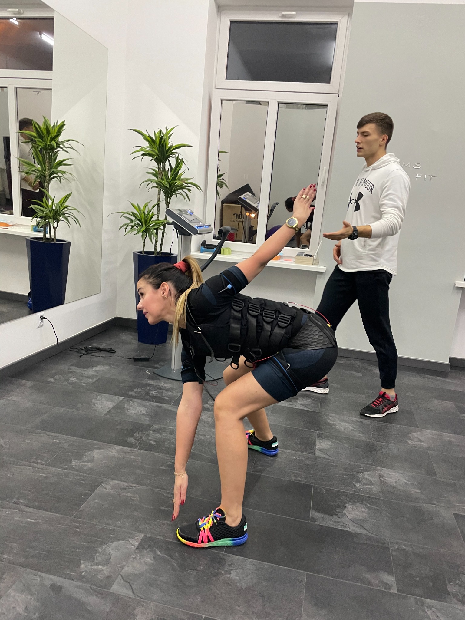 Kobieta w kombinezonie EMS wykonuje ćwiczenie pod okiem trenera personalnego w jasnym wnętrzu siłowni, widoczne rośliny w donicach.