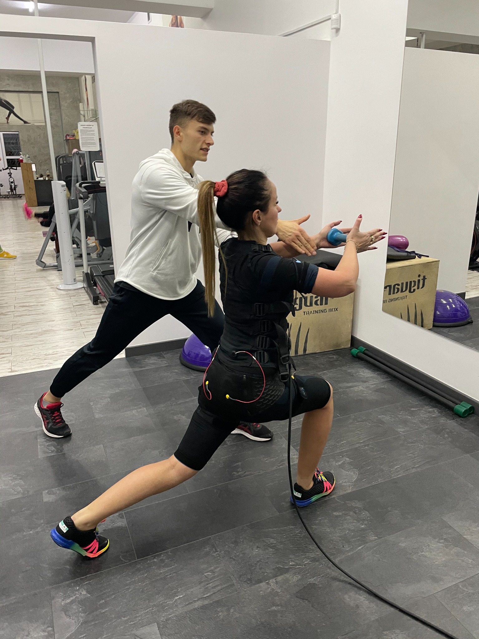 Kobieta w stroju do elektrostymulacji mięśni wykonuje wykrok pod nadzorem mężczyzny w jasnej bluzie w siłowni.