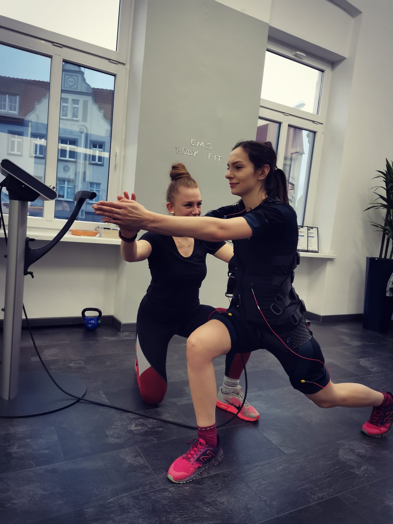 Kobieta w kombinezonie EMS ćwiczy pod okiem instruktorki w jasnym pomieszczeniu z dużym oknem, na ścianie napis 'EMS Body Fit'.
