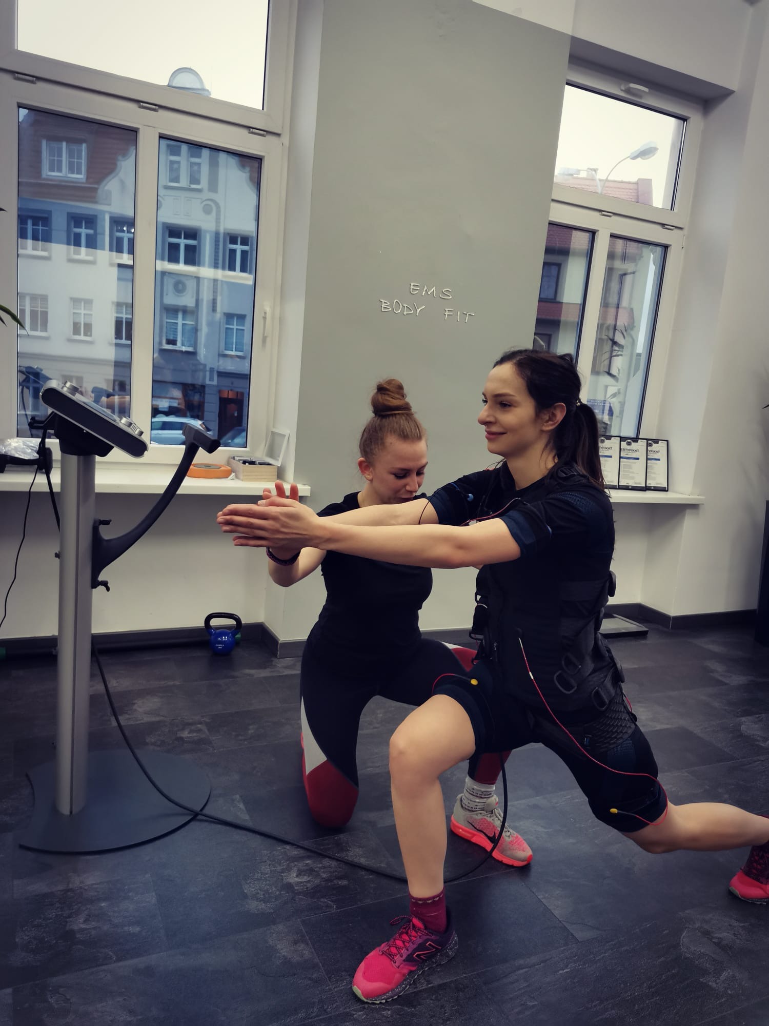 Kobieta w stroju do elektrostymulacji EMS wykonuje wykrok pod nadzorem instruktorki w studiu z dużymi oknami i napisem 'EMS BODY FIT' na ścianie.