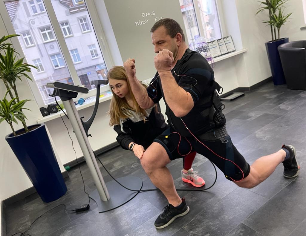 Mężczyzna w kombinezonie EMS wykonuje wypad w przód pod nadzorem kobiety w jasnym wnętrzu siłowni.