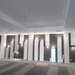 Wnętrze biura w trakcie wykończenia, ściana pomalowana na ciemny kolor z białymi, pionowymi elementami dekoracyjnymi i oświetleniem punktowym, widoczne kable wystające z sufitu, podłoga...