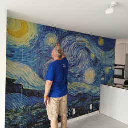Artur Szostak - Mężczyzna w niebieskiej koszulce i beżowych szortach przykłada dłoń do ściany z tapetą przedstawiającą motyw 'Gwiaździstej Nocy' Van Gogha w jasnym wnętrzu.