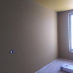 Artur Szostak - Pomalowane na beżowo wnętrze pokoju z oknem i żółtym sufitem, widoczne gniazdka elektryczne na ścianie oraz opakowanie paneli na podłodze.