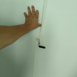 Artur Szostak - Nakładanie jasnej tapety na ścianę, widoczna dłoń dociskająca brzeg tapety przy listwie mocującej.