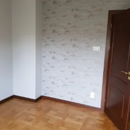 Wykończone wnętrze pokoju: gładkie, szare ściany z delikatnym, nieregularnym wzorem w odcieniach brązu, drewniana podłoga w parkiet, ciemnobrązowe listwy przypodłogowe i drzwi z złotą klamką.