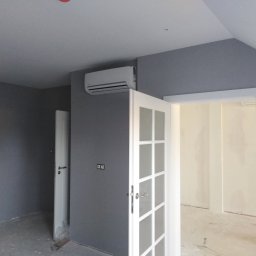 Artur Szostak - Pokój po malowaniu ścian na szaro, widoczny klimatyzator, białe drzwi z przeszkleniami i fragment podłogi w trakcie remontu.