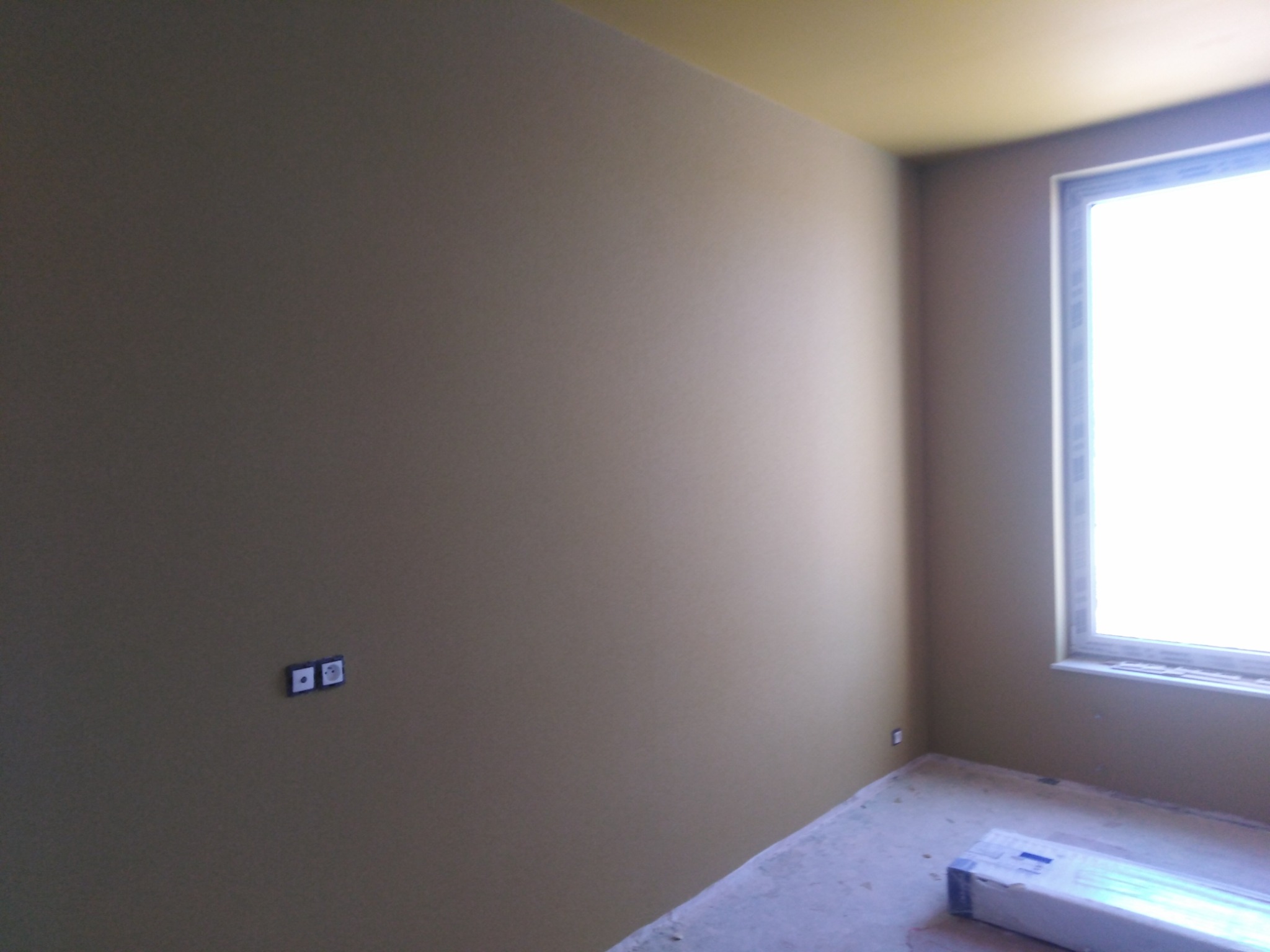 Pomalowane na beżowo wnętrze pokoju z oknem i żółtym sufitem, widoczne gniazdka elektryczne na ścianie oraz opakowanie paneli na podłodze.