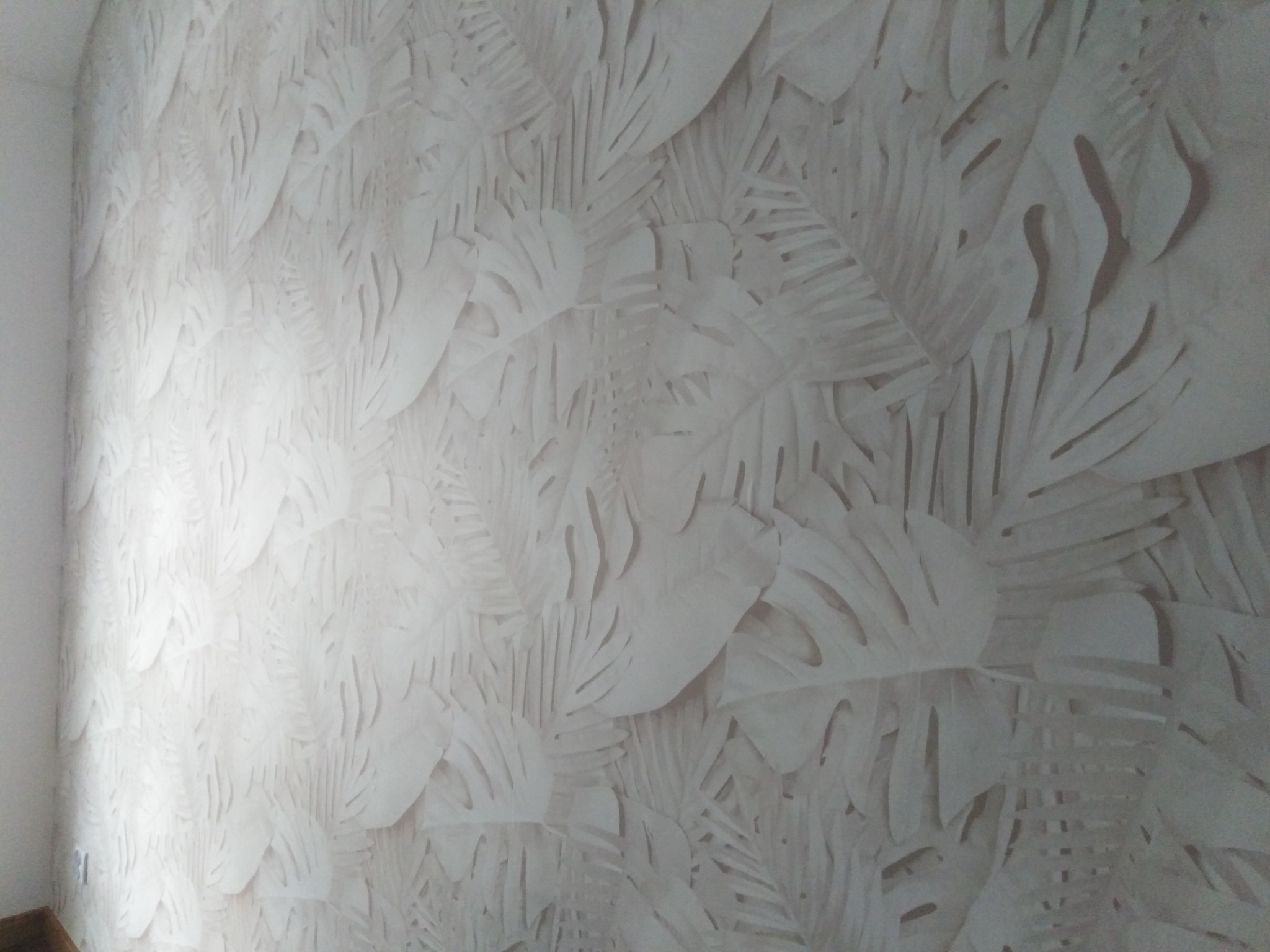 Ściana pokryta tapetą z tłoczonym wzorem liści monstery i palm w odcieniach bieli i beżu, z delikatnym cieniowaniem podkreślającym trójwymiarowość wzoru.