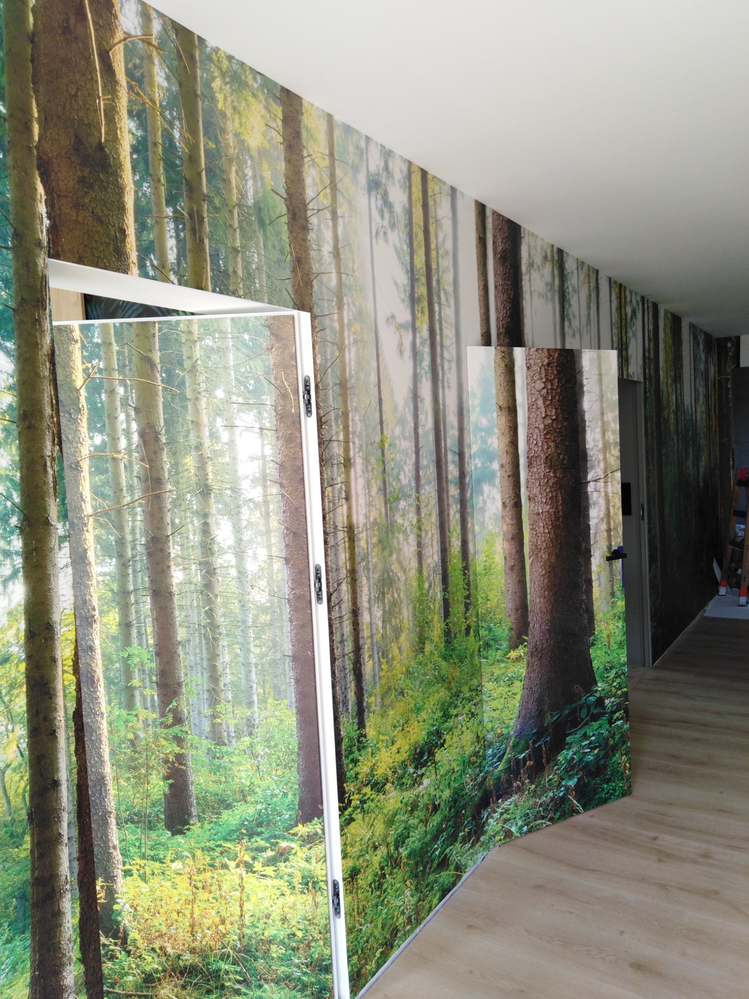 Ściana z fototapetą przedstawiającą las iglasty, widoczna krawędź fototapety na ścianie i na skrzydle drzwiowym, jasna podłoga panelowa.