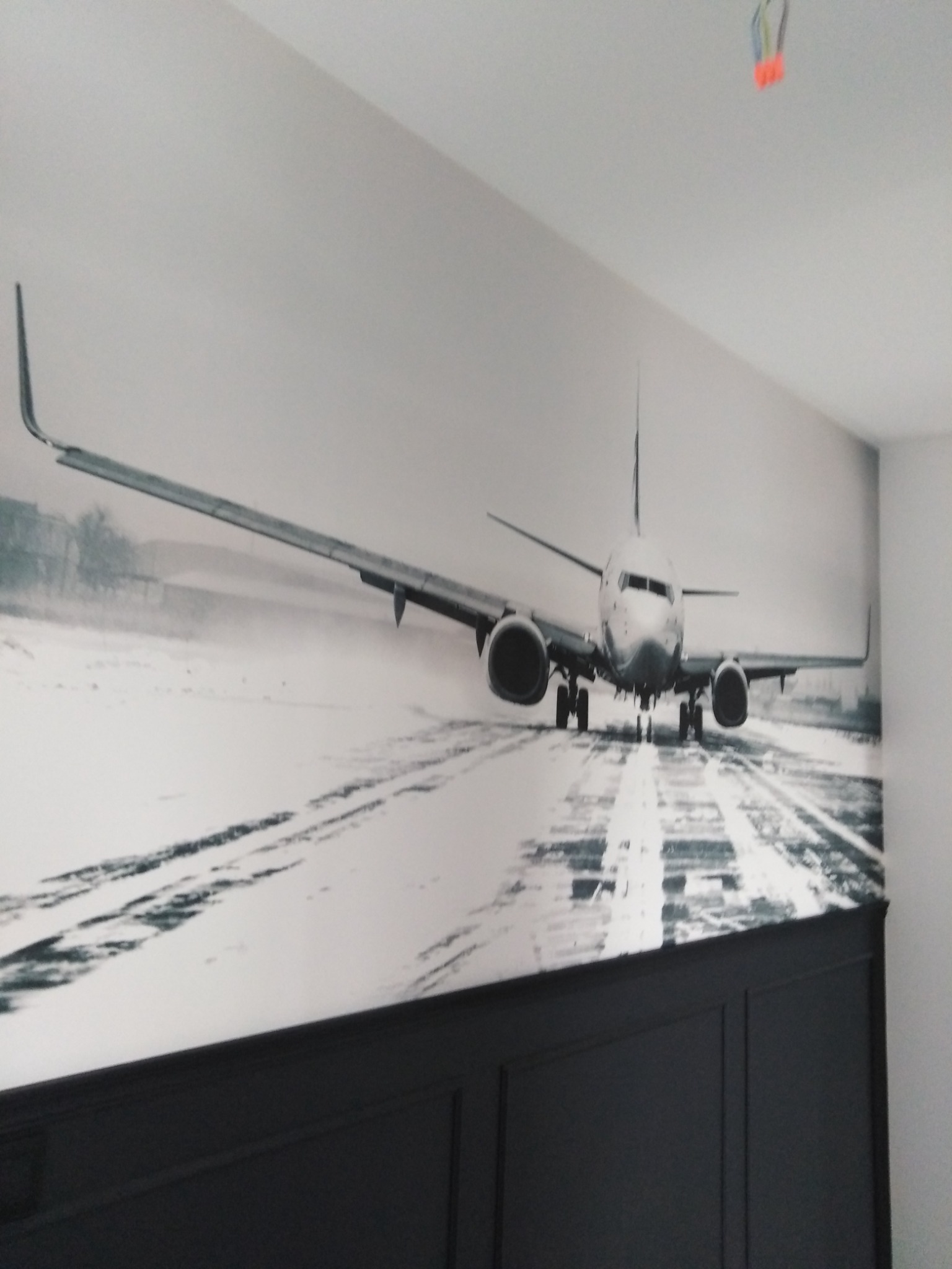 Czarno-biała fototapeta przedstawiająca samolot na pasie startowym, zamontowana nad ciemną, panelową ścianą.