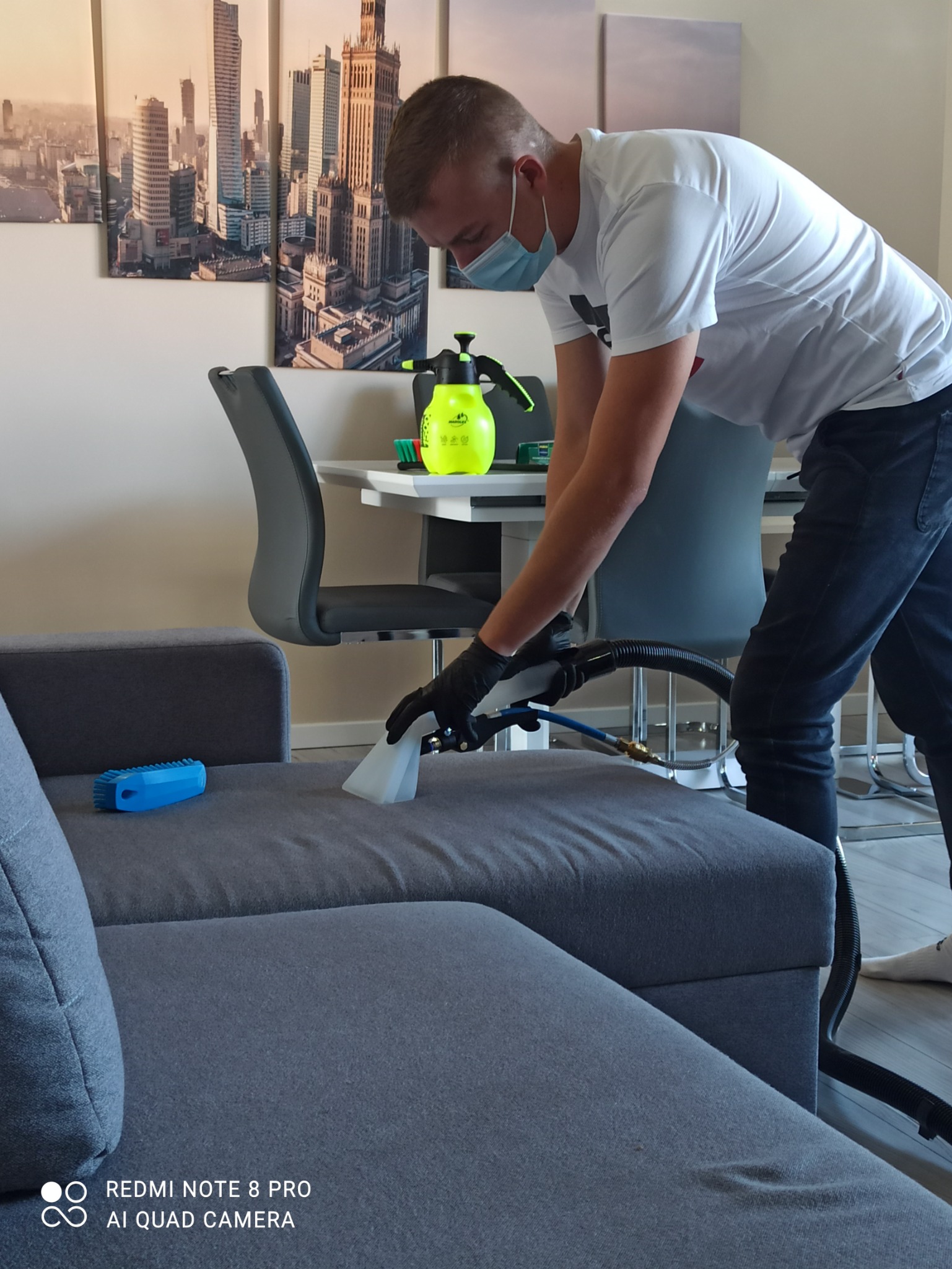 Mężczyzna w masce i rękawicach czyszczący szarą, narożną kanapę za pomocą odkurzacza piorącego. Na kanapie leży niebieska szczotka. W tle widoczny stół z krzesłami i obraz z panoramą miasta.