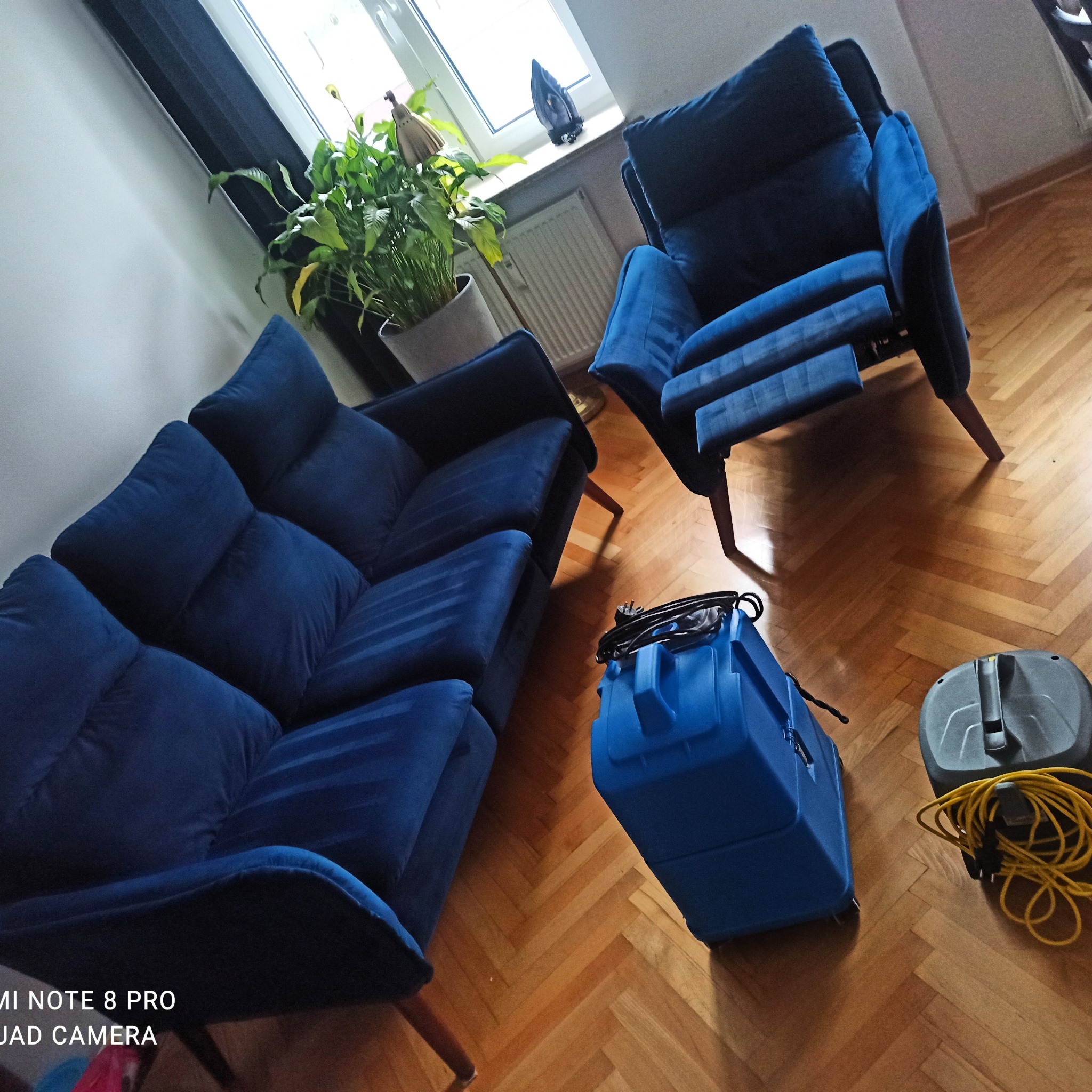 Niebieski zestaw wypoczynkowy (sofa i fotel) na parkiecie, obok niebieski i szary sprzęt do czyszczenia tapicerki z żółtym kablem, w tle roślina doniczkowa i okno.
