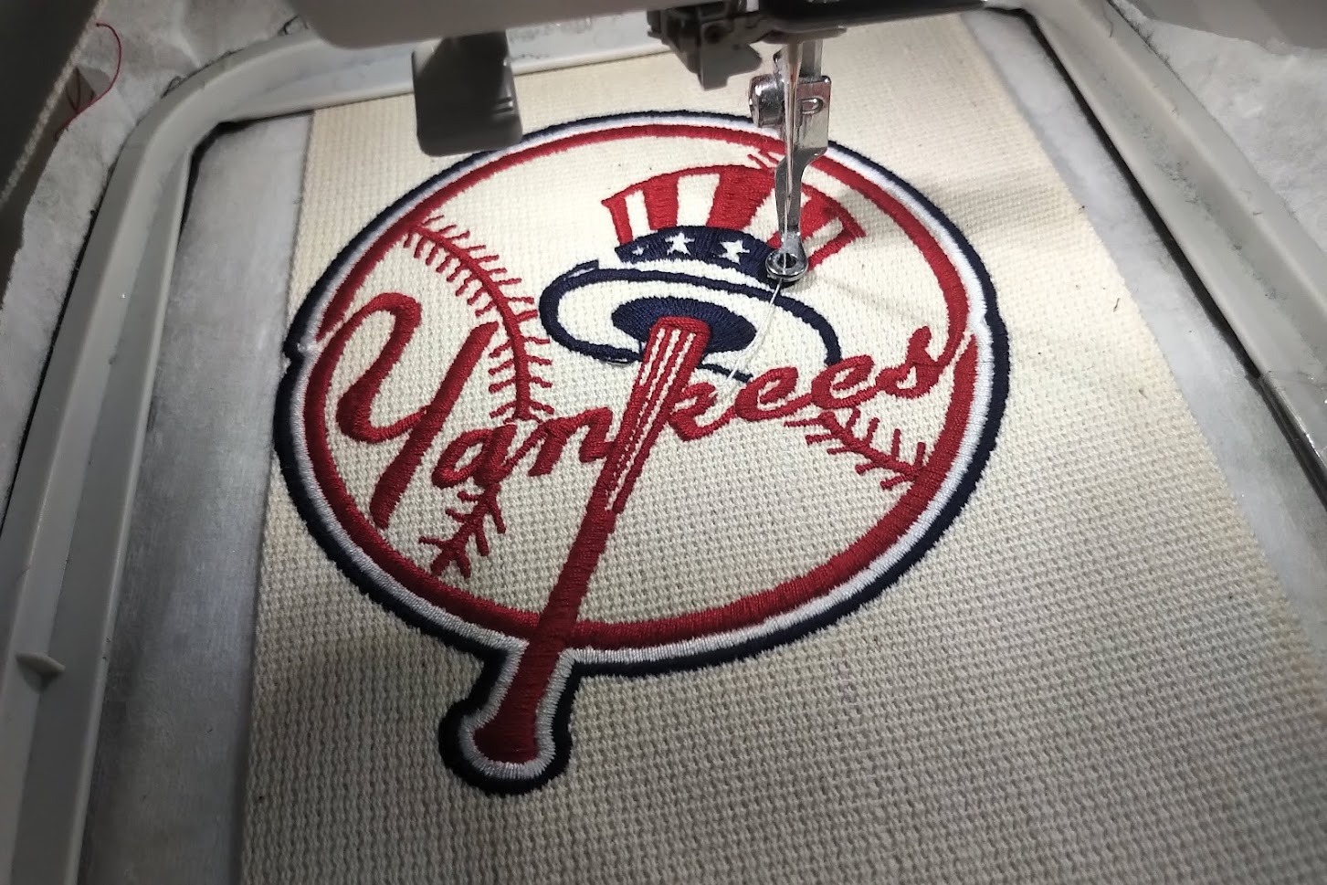 Automatyczna hafciarka w trakcie wykonywania kolorowego haftu z motywem baseballowym na kremowym materiale, widoczny detal igły i napięty materiał w tamborku.