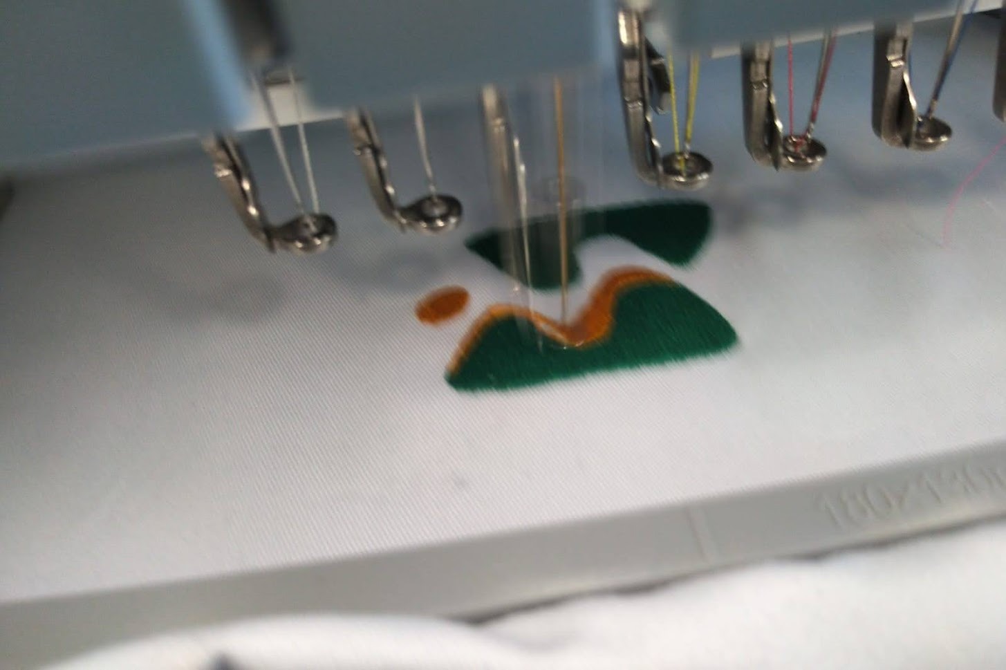 Igły maszyny haftującej zielono-pomarańczowy wzór na białym materiale, zbliżenie na proces tworzenia haftu.