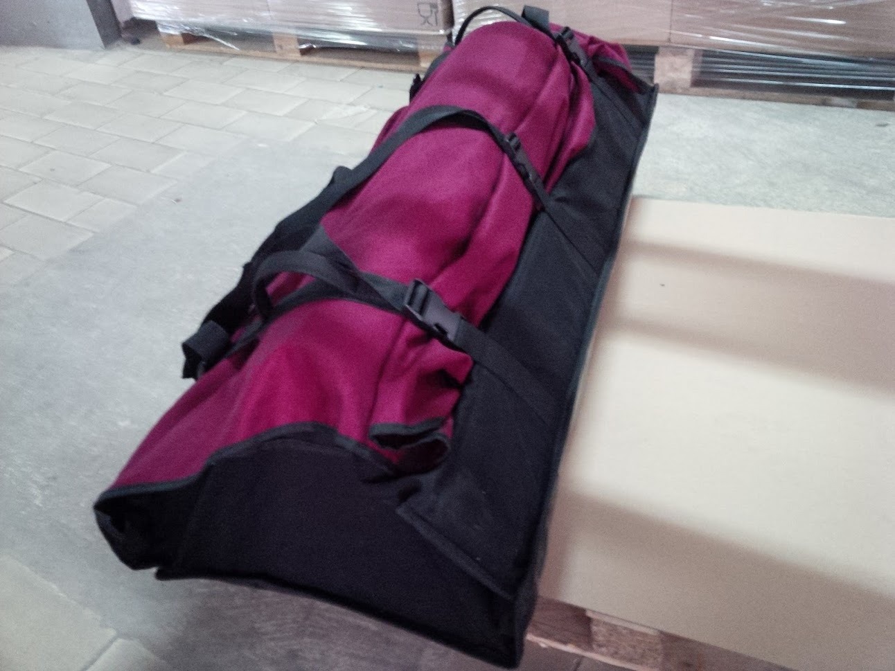 Podłużny, bordowy pokrowiec transportowy z czarnymi wstawkami i paskami, leżący na palecie w magazynie.