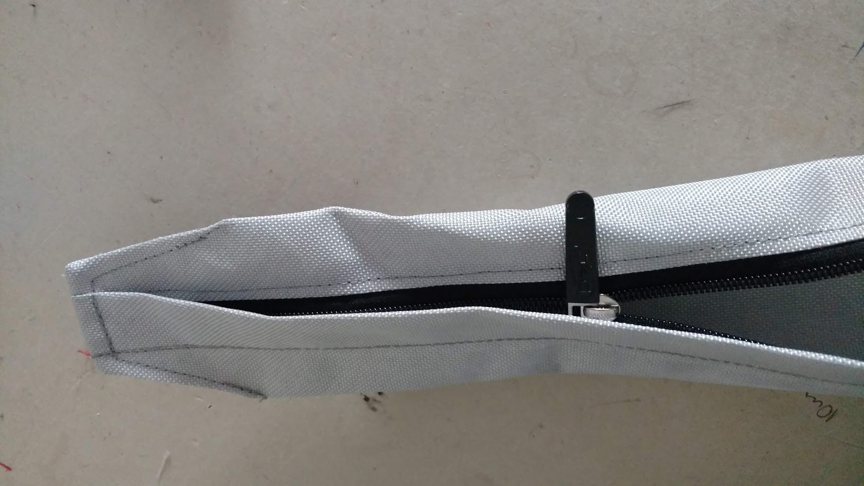 Fragment szarego pokrowca z zamkiem błyskawicznym i czarnym suwakiem, widoczne szwy i fragment materiału.