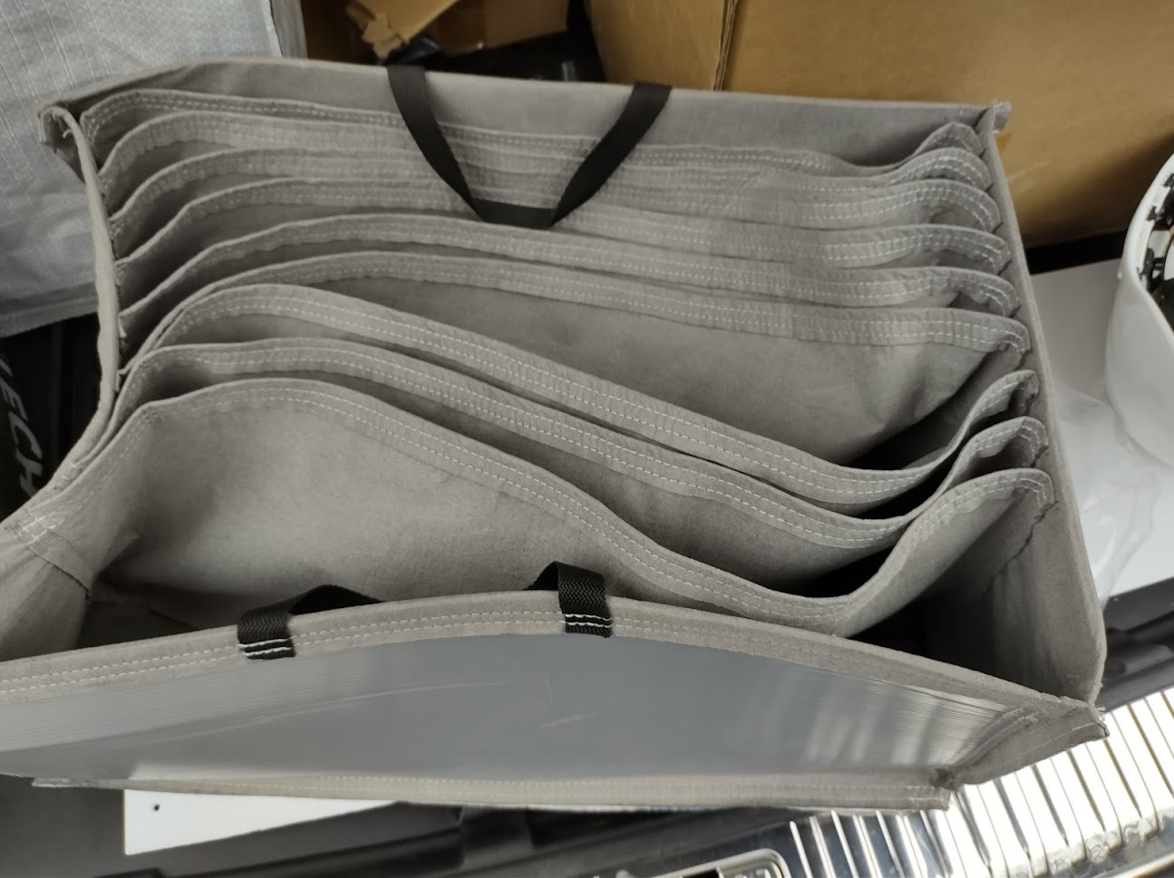 Stos szarych, płaskich pokrowców z uchwytami, widoczne szwy i plastikowe wzmocnienie krawędzi.