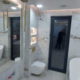 ProGres Tiler - Nowoczesna łazienka z marmurowymi płytkami, złotymi lampami wiszącymi i czarną ramą drzwiową, odbicie osoby w drzwiach.
