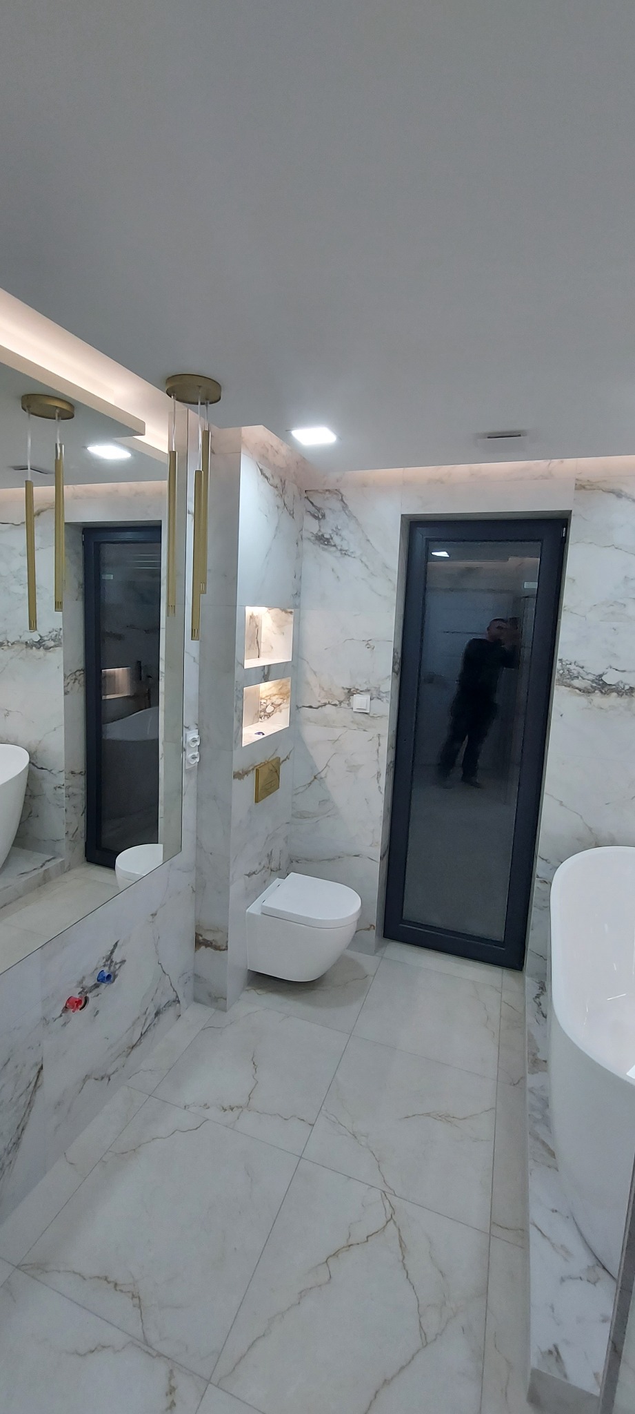 Nowoczesna łazienka z marmurowymi płytkami, złotymi lampami wiszącymi i czarną ramą drzwiową, odbicie osoby w drzwiach.