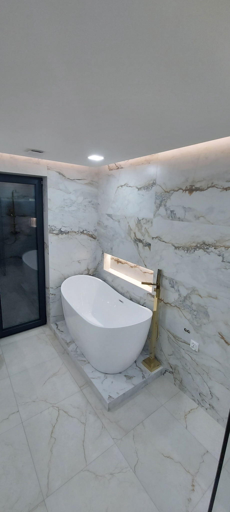 Nowoczesna łazienka z wolnostojącą wanną, złotą armaturą i marmurowymi płytkami na ścianach i podłodze, oświetlona wnęka nad wanną.