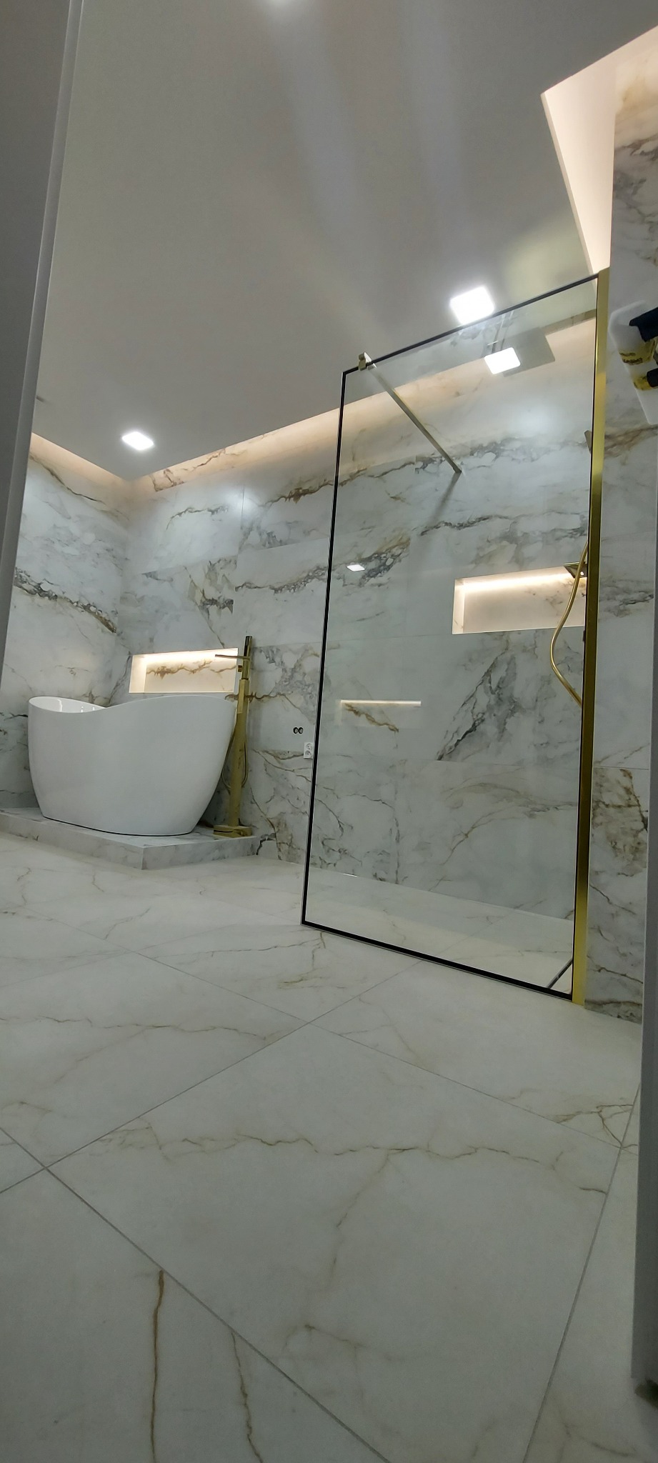 Nowoczesna łazienka z marmurowymi płytkami, wolnostojącą wanną i kabiną prysznicową ze złotymi detalami, oświetlona subtelnym światłem.