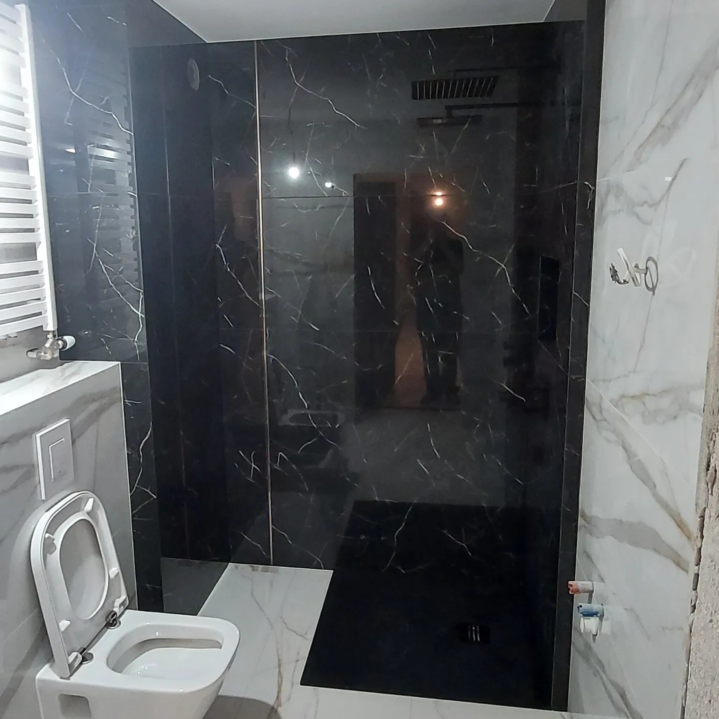 Nowoczesna łazienka z czarnymi marmurowymi płytkami w strefie prysznica i białymi marmurowymi płytkami na pozostałych ścianach, widoczna toaleta z otwartą klapą, grzejnik drabinkowy oraz czarny...