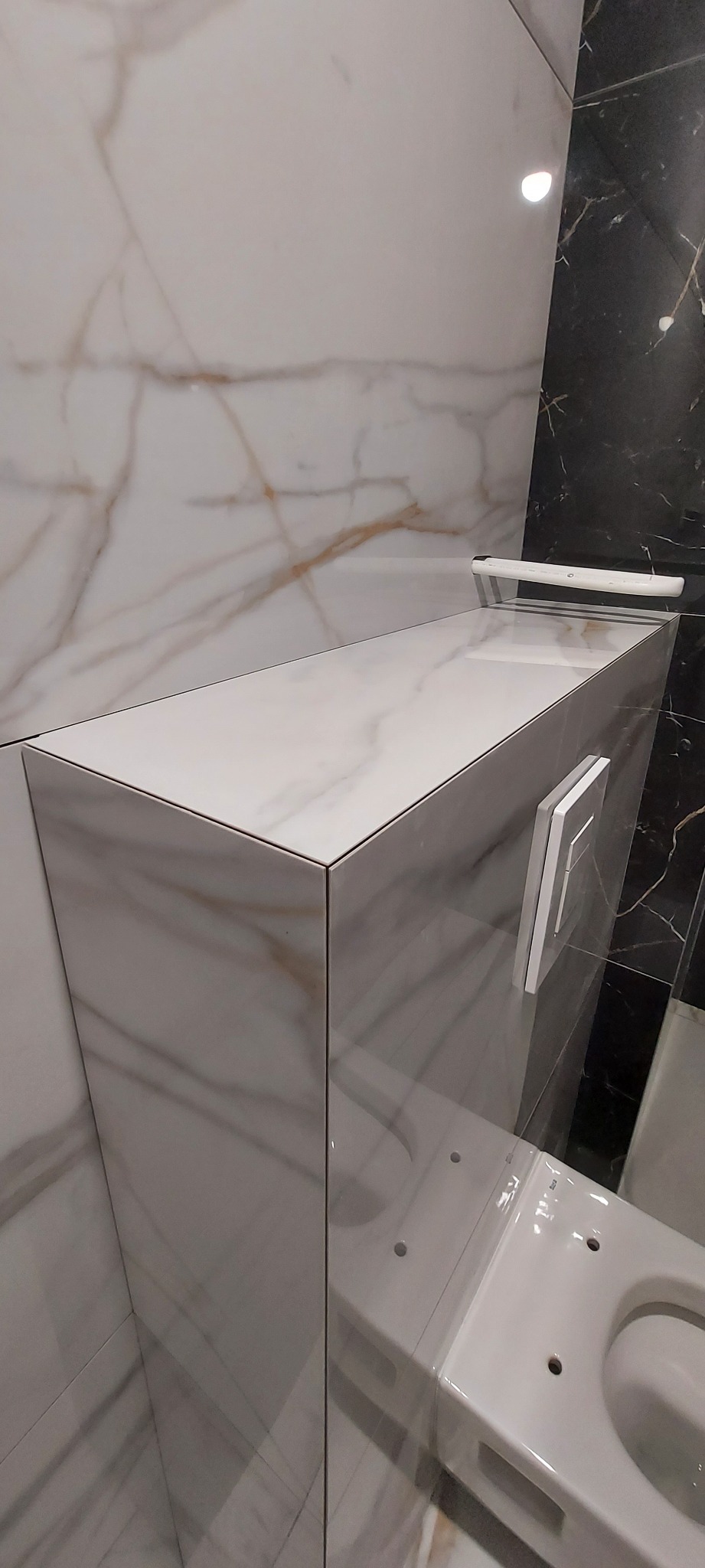 Obudowa stelaża podtynkowego WC wykończona płytkami imitującymi biały marmur z delikatnym złotym żyłowaniem, pasująca do okładziny ścian, widoczny przycisk spłukiwania i fragment miski WC