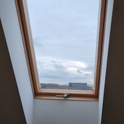 Lux-bud - Okno dachowe z drewnianą ramą, widok na pochmurne niebo i odległe budynki, widoczny mechanizm otwierania.