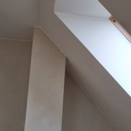 Lux-bud - Ściana poddasza z oknem dachowym po nałożeniu gładzi, widoczny fragment drewnianego elementu okna i puszka elektryczna w ścianie.