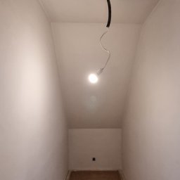 Lux-bud - Wnętrze małego pomieszczenia z gładkimi, pomalowanymi na biało ścianami, z widocznym kablem z żarówką zwisającym z sufitu i surową podłogą przygotowaną do dalszych prac wykończeniowych.