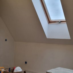 Lux-bud - Poddasze z oknem dachowym po nałożeniu gładzi, widoczne narzędzia i akcesoria malarskie, w tym wiadro farby i odkurzacz przemysłowy.