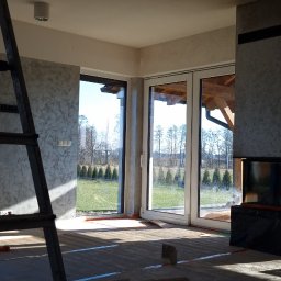 Lux-bud - Przygotowanie do ułożenia podłogi w jasnym salonie w Oleśnicy, widoczne rolki materiału izolacyjnego, drabina i duże okna z widokiem na zewnątrz.