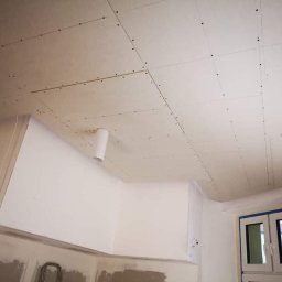 Lux-bud - Sufit z zamontowanymi płytami, widoczne ślady po wkrętach, białe ściany i rura wentylacyjna, pomieszczenie w trakcie prac wykończeniowych.