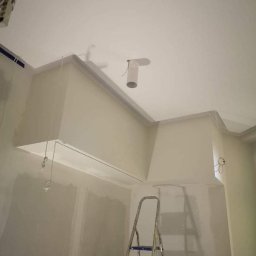 Lux-bud - Wnętrze w trakcie remontu: ściany pokryte gładzią, widoczna drabina aluminiowa, wystające przewody elektryczne, sufit z elementami zabudowy gipsowo-kartonowej i otworem wentylacyjnym.