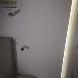 Lux-bud - Wnętrze z gładką, białą powierzchnią sufitu, oświetleniem LED wzdłuż krawędzi i wystającymi przewodami elektrycznymi oraz widocznym fragmentem ściany.