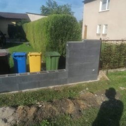 Lux-bud - Wznoszenie niskiego murku z szarych bloczków betonowych na trawniku, widoczne trzy kolorowe pojemniki na śmieci w tle oraz cień osoby trzymającej poziomicę.