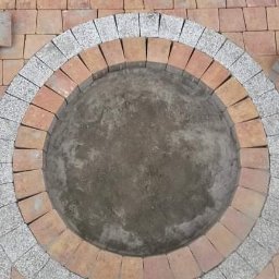 Lux-bud - Budowa okrągłego paleniska z cegieł i kostki granitowej, widoczne buty robotnika i drewniana listwa poziomująca.