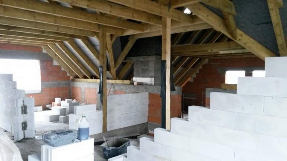 Surowy stan poddasza z widoczną konstrukcją drewnianą dachu, murem z czerwonej cegły, ścianami wznoszonymi z białych bloczków, narzędziami i materiałami budowlanymi.