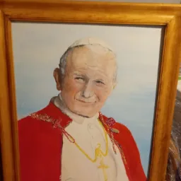 Portret Jana Pawła II w złotej ramie, malowany farbami olejnymi, z widocznymi pociągnięciami pędzla, ustawiony na wzorzystym materiale.