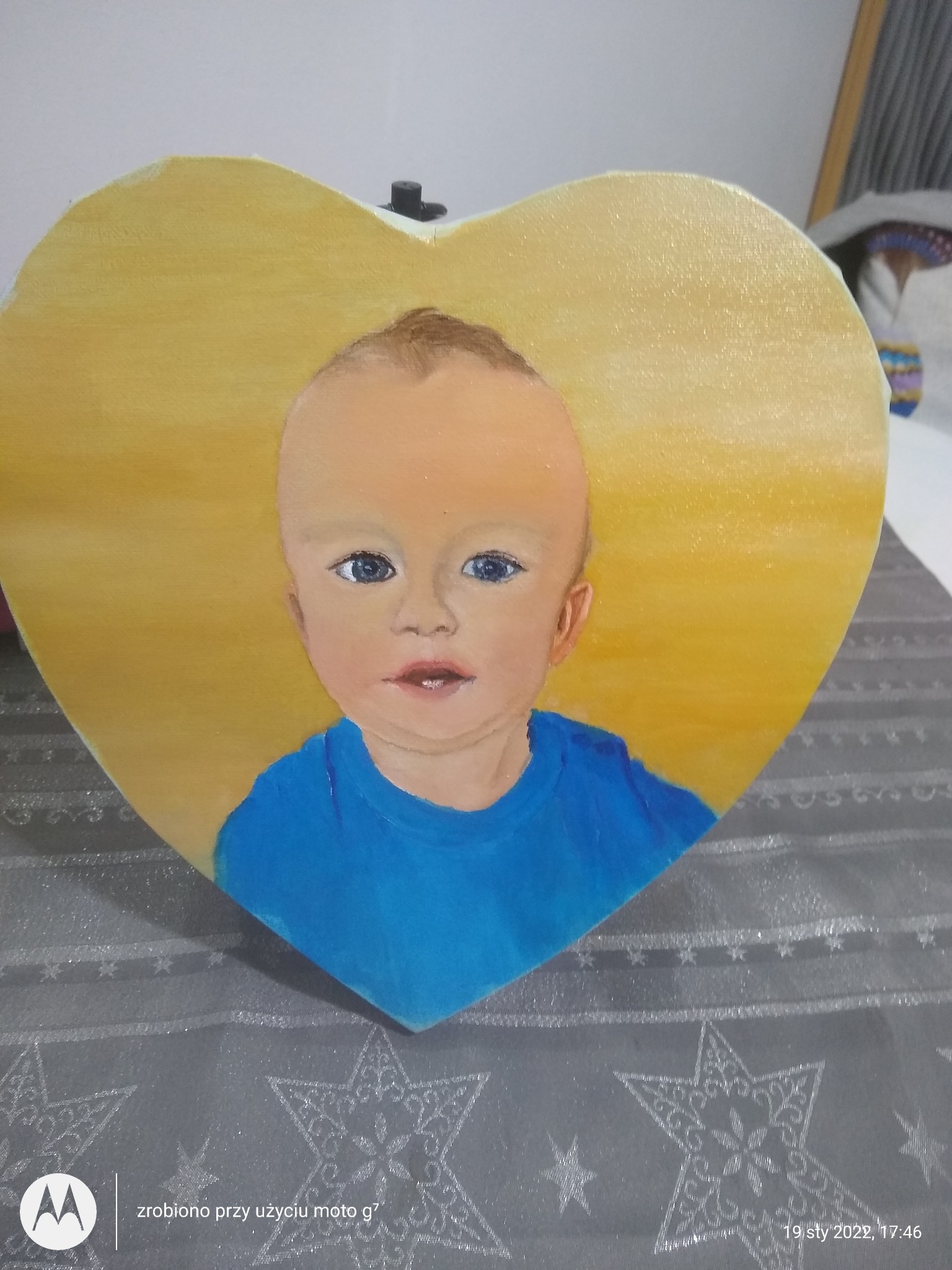 Portret dziecka w niebieskiej koszulce, namalowany na drewnianym sercu z żółtym tłem, leżący na szarym obrusie w gwiazdy.